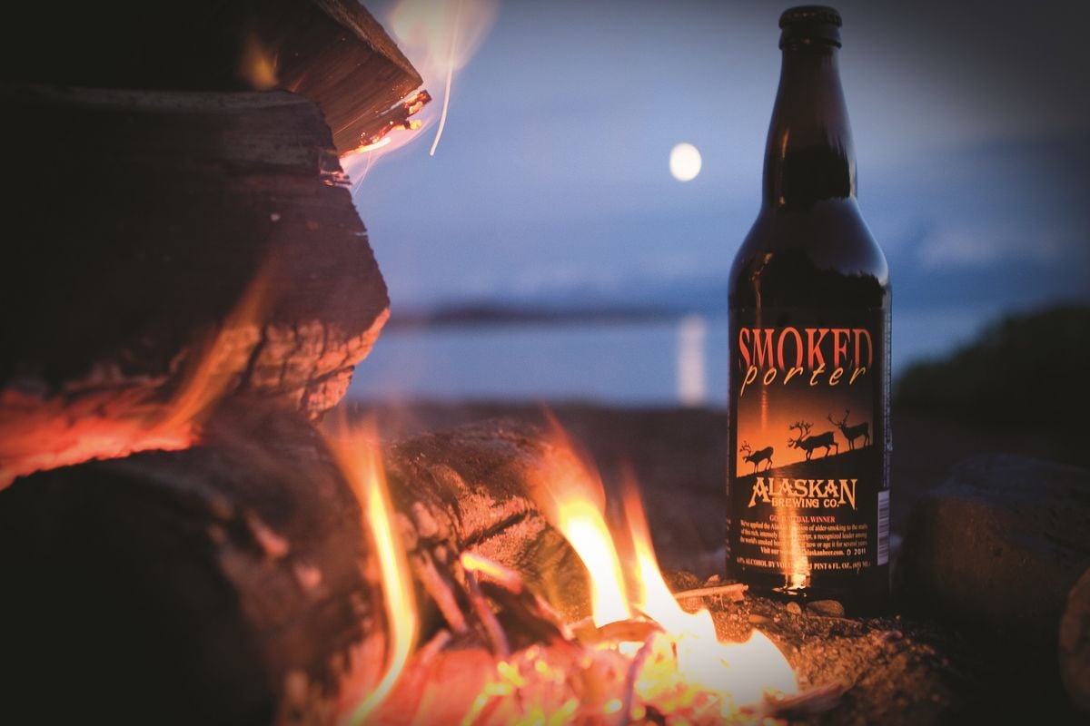 Photo of a bottle of Smoked Porter by Alaskan Brewing Co. placed by a fire by a lake in the evening.